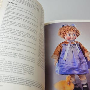 Pode incluir: Uma boneca vintage com cabelo loiro encaracolado usando um vestido azul e um chap&eacute;u branco. A boneca est&aacute; segurando um pintinho amarelo em sua m&atilde;o. A boneca est&aacute; representada em um fundo branco.