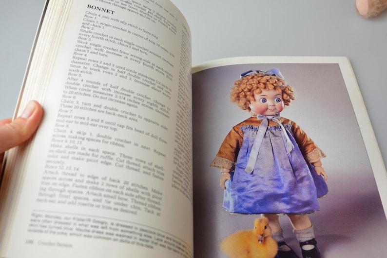 Pode incluir: Uma boneca vintage com cabelo loiro encaracolado usando um vestido azul e um chap&eacute;u branco. A boneca est&aacute; segurando um pintinho amarelo em sua m&atilde;o. A boneca est&aacute; representada em um fundo branco.