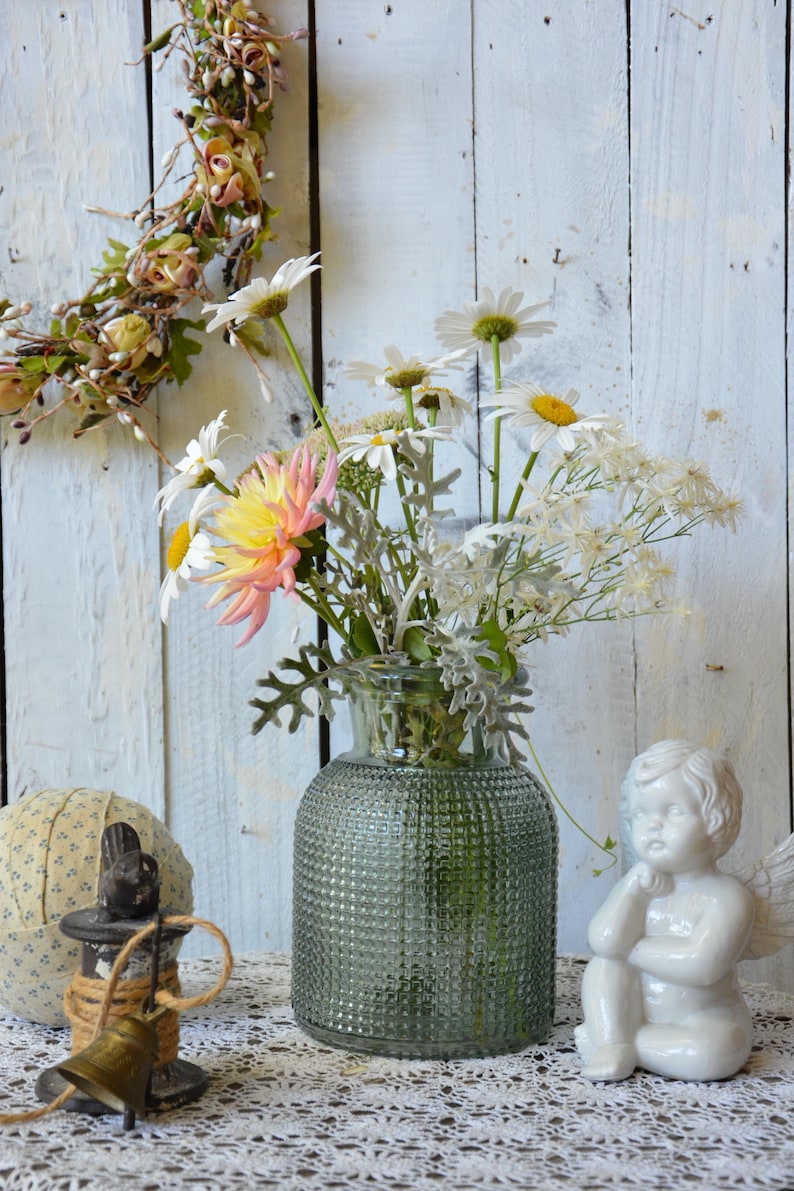 May include: A clear green glass vase with a textured hobnail pattern holds a bouquet of white daisies, a pink and yellow dahlia, and silver-toned dusty miller foliage. The vase sits alongside a small white ceramic angel figurine and a metal bell.