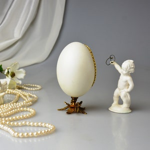 Puede incluir: Un huevo decorativo blanco con un borde dorado se asienta sobre un soporte dorado de tres patas. Una peque&ntilde;a figurita de porcelana blanca de un querub&iacute;n se encuentra a la derecha del huevo.