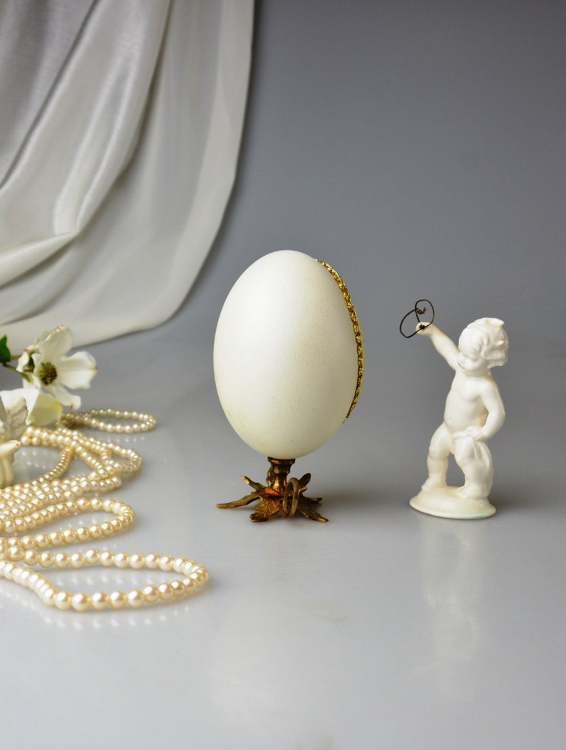 Puede incluir: Un huevo decorativo blanco con un borde dorado se asienta sobre un soporte dorado de tres patas. Una peque&ntilde;a figurita de porcelana blanca de un querub&iacute;n se encuentra a la derecha del huevo.