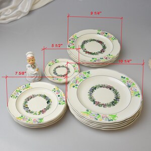 May include: A collection of cream-colored ceramic plates with floral wreath designs. The plates vary in size, with the largest measuring 10 1/4 inches in diameter and the smallest 5 1/2 inches. A decorative figurine is also present.