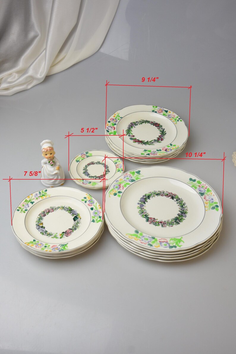 May include: A collection of cream-colored ceramic plates with floral wreath designs. The plates vary in size, with the largest measuring 10 1/4 inches in diameter and the smallest 5 1/2 inches. A decorative figurine is also present.