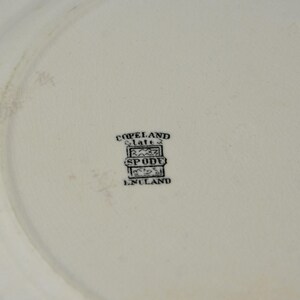 May include: Close-up of a white plate with a faded design. The plate has a black stamp that reads "Copeland Spode England". The plate is likely vintage or antique.