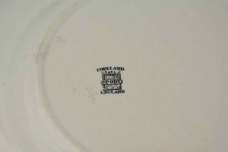 May include: Close-up of a white plate with a faded design. The plate has a black stamp that reads "Copeland Spode England". The plate is likely vintage or antique.