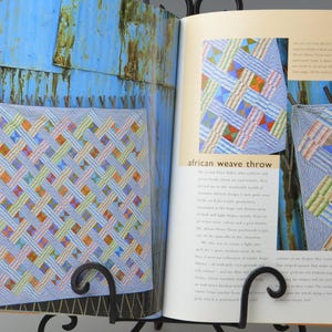 May include: An open book showing a colourful quilt with a woven design, labelled "African Weave Throw." The quilt has a light blue base with interwoven strips of various colours. The book is supported by a black metal stand.