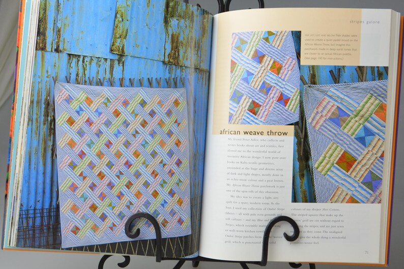 May include: An open book showing a colourful quilt with a woven design, labelled "African Weave Throw." The quilt has a light blue base with interwoven strips of various colours. The book is supported by a black metal stand.