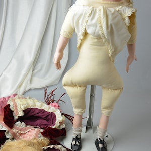 May include: A porcelain doll with a pale complexion and blue eyes, wearing a white dress with lace trim and beige pants. The doll is standing on a metal stand and has a wig of blonde hair.