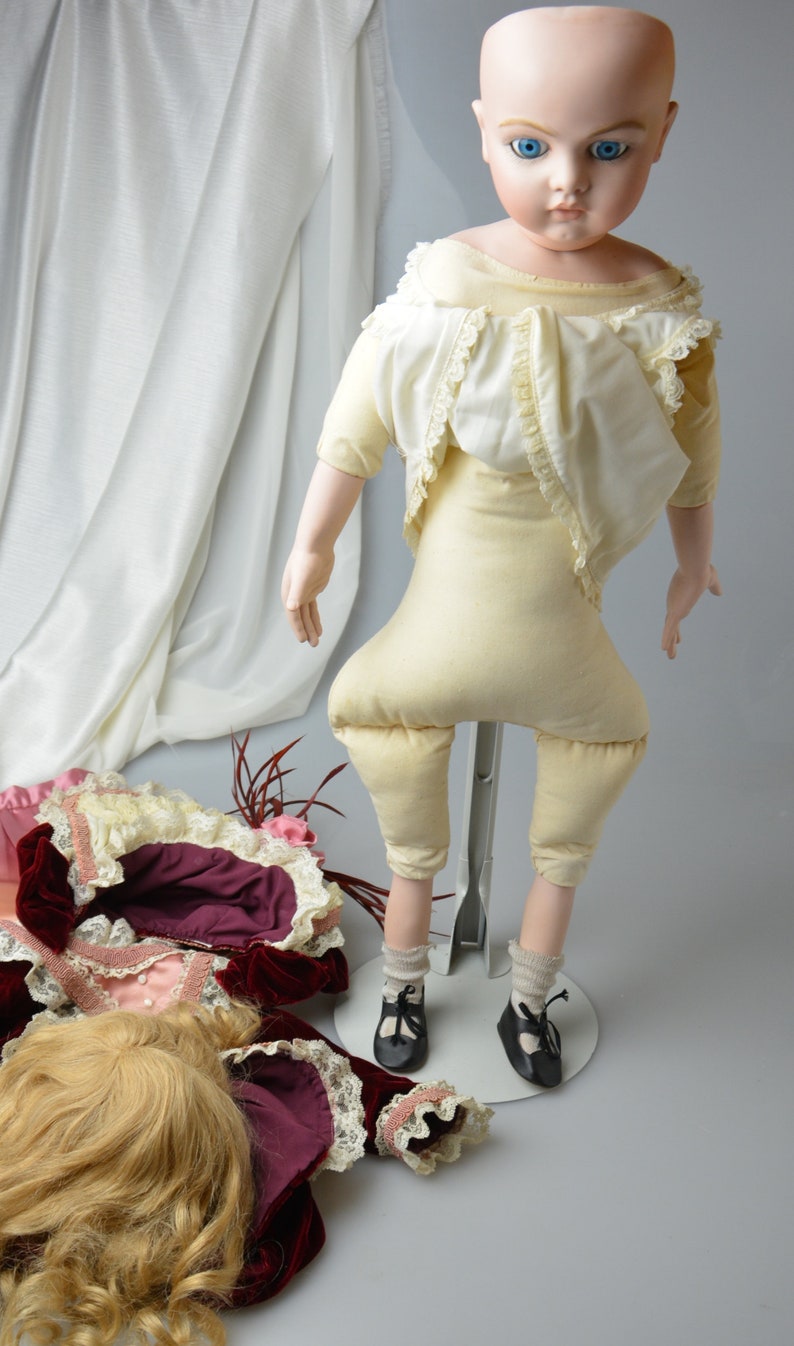 May include: A porcelain doll with a pale complexion and blue eyes, wearing a white dress with lace trim and beige pants. The doll is standing on a metal stand and has a wig of blonde hair.