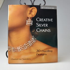 May include: A book cover for "Creative Silver Chains" by Chantal Lise Saunders. The cover features a woman wearing silver earrings and a necklace. The book is displayed in a decorative black metal stand.