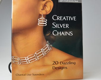 May include: A book cover for "Creative Silver Chains" by Chantal Lise Saunders. The cover features a woman wearing silver earrings and a necklace. The book is displayed in a decorative black metal stand.