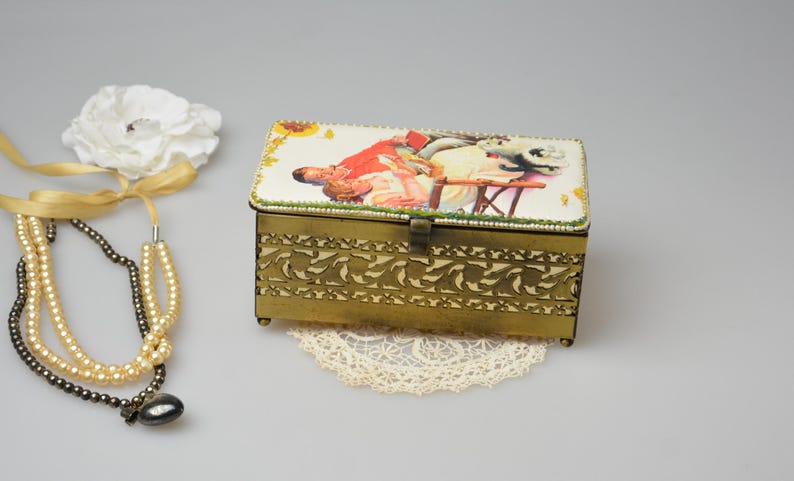 May include: A vintage brass jewelry box with a hinged lid, featuring a decorative floral design on the sides and a painted scene on the top. A white flower, gold ribbon, and pearl necklace are next to the box.