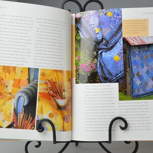 May include: An open book on a black metal stand displays colourful images. One page shows a yellow and orange patterned fabric on a chair. The other page features a blue quilt and a blue and white patterned structure.