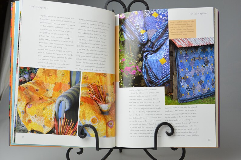 May include: An open book on a black metal stand displays colourful images. One page shows a yellow and orange patterned fabric on a chair. The other page features a blue quilt and a blue and white patterned structure.