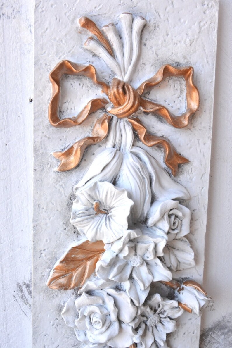 May include: A white plaster wall decor with a gold ribbon bow and white flowers. The flowers are in a cluster and have a 3D effect.