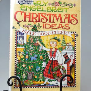 Pode incluir: Um livro intitulado "Mary Engelbreit Christmas Ideas" exibido em um cavalete de metal preto. A capa apresenta uma ilustração festiva de uma menina decorando uma árvore de Natal, com o texto "Make Good Cheer!"