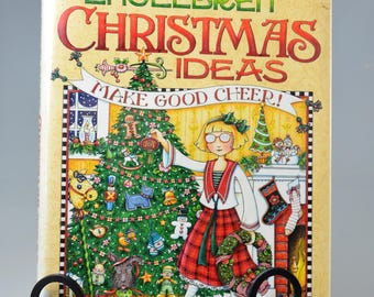 Puede incluir: Un libro titulado "Mary Engelbreit Christmas Ideas" exhibido en un caballete de metal negro. La portada presenta una ilustración festiva de una niña decorando un árbol de Navidad, con el texto "Make Good Cheer!"