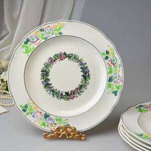 May include: White ceramic plates with a floral wreath design in the center and floral accents around the rim. The plates have a scalloped edge and a thin black border. A stack of plates is visible.