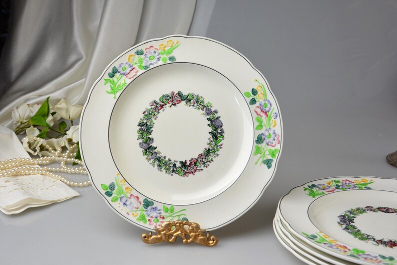 May include: White ceramic plates with a floral wreath design in the center and floral accents around the rim. The plates have a scalloped edge and a thin black border. A stack of plates is visible.