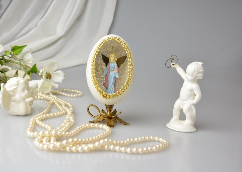 Puede incluir: Un huevo de porcelana blanca con un borde dorado y una figurita de &aacute;ngel en miniatura en su interior. El huevo est&aacute; sobre un soporte dorado con un dise&ntilde;o de hojas. Tambi&eacute;n hay un collar de perlas y una figurita de querub&iacute;n de porcelana blanca en la imagen.