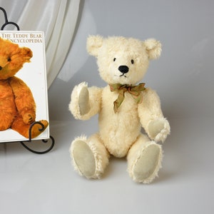 May include: A white teddy bear with a green bow tie is sitting on a white surface. The teddy bear has black eyes and a black nose. The teddy bear is wearing a green bow tie. The teddy bear is sitting on a white surface.