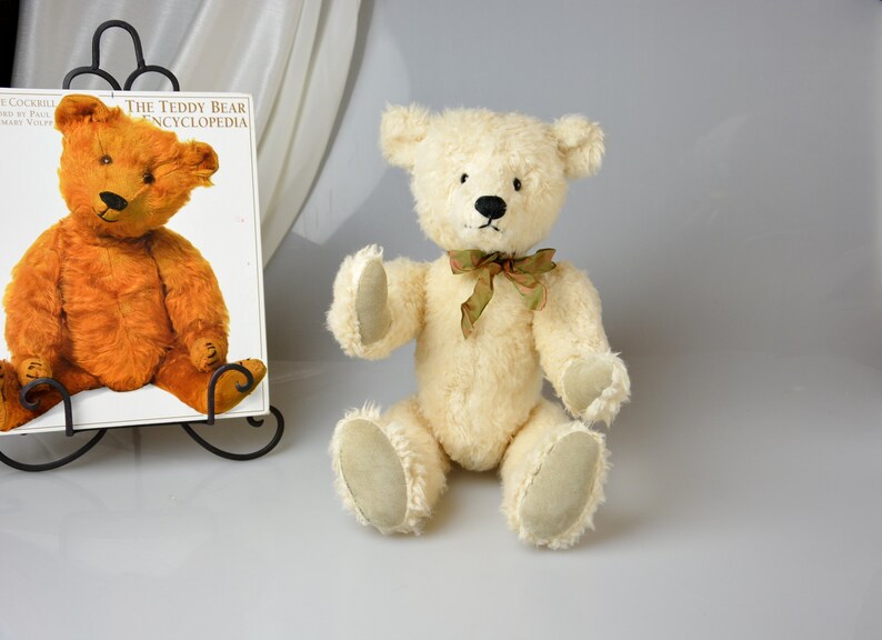 May include: A white teddy bear with a green bow tie is sitting on a white surface. The teddy bear has black eyes and a black nose. The teddy bear is wearing a green bow tie. The teddy bear is sitting on a white surface.