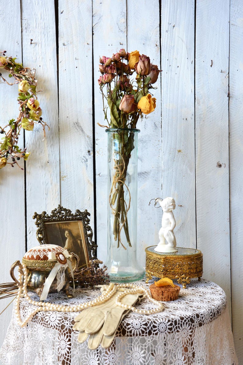 K&ouml;nnte beinhalten: Ein Stillleben mit einer transparenten Glasvase mit getrockneten Rosen, einem gerahmten Bild, einer dekorativen Box, einem Nadelkissen, einer Perlenkette und Handschuhen. Die Gegenst&auml;nde sind auf einem runden Tisch mit einer wei&szlig;en Spitzentischdecke angeordnet.