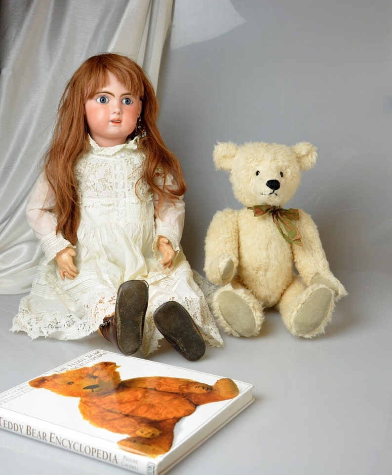 May include: A porcelain doll with long brown hair wearing a white lace dress sits next to a white teddy bear with a green bow. A book titled "The Teddy Bear Encyclopedia" is in the foreground.