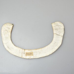 May include: A white fabric collar with a small label attached. The collar is shaped like a crescent moon and is made of a soft, pliable material.