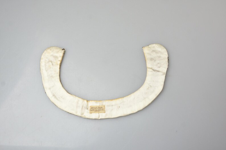 May include: A white fabric collar with a small label attached. The collar is shaped like a crescent moon and is made of a soft, pliable material.