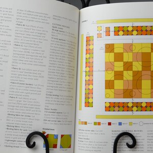May include: Open book showing a quilt design diagram with yellow, orange, and brown squares and circles. Text on the left page details the quilt's fabrics and construction. The book is held open by a black metal stand. Measurements are in cm.