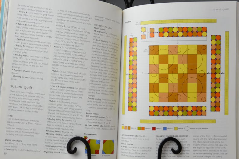 May include: Open book showing a quilt design diagram with yellow, orange, and brown squares and circles. Text on the left page details the quilt's fabrics and construction. The book is held open by a black metal stand. Measurements are in cm.