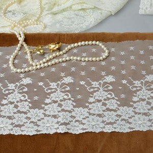 May include: White lace trim with a floral pattern. The lace is delicate and has a scalloped edge. It is shown on a brown surface with a pearl necklace.