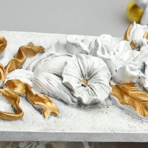 May include: White and gold decorative wall plaque with a floral design. The plaque features a large bow, flowers, and leaves. The flowers are white and the leaves are gold.