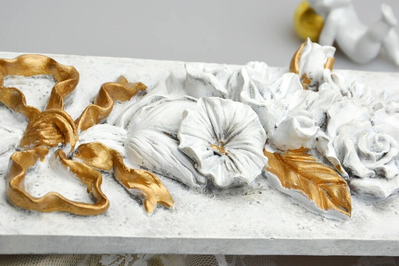 May include: White and gold decorative wall plaque with a floral design. The plaque features a large bow, flowers, and leaves. The flowers are white and the leaves are gold.