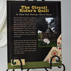Może przedstawiać: Okładka książki "The Circuit Rider's Quilt" ma czarne tło z złotym tekstem. Tytuł to "The Circuit Rider's Quilt", a podtytuł to "An Album Quilt Honoring a Beloved Minister". Okładka zawiera r&oacute;wnież zdjęcie małego kamiennego budynku z kocem wiszącym na krześle przed nim.