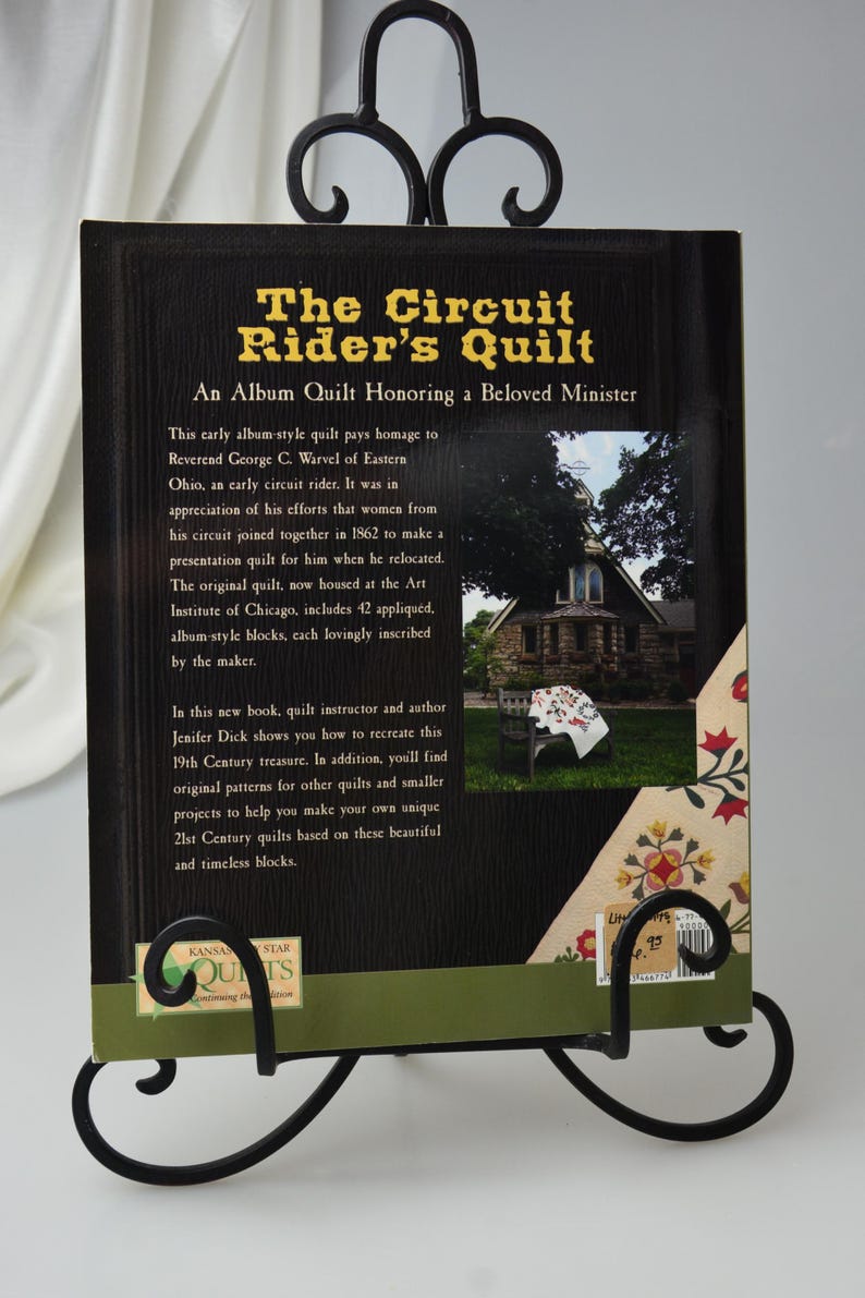 Może przedstawiać: Okładka książki "The Circuit Rider's Quilt" ma czarne tło z złotym tekstem. Tytuł to "The Circuit Rider's Quilt", a podtytuł to "An Album Quilt Honoring a Beloved Minister". Okładka zawiera r&oacute;wnież zdjęcie małego kamiennego budynku z kocem wiszącym na krześle przed nim.