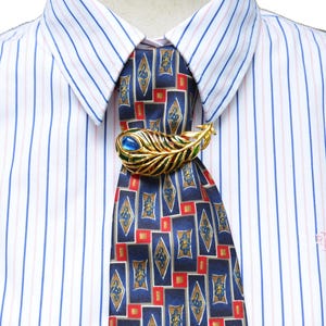 May include: A gold-toned metal brooch shaped like a peacock feather with blue and green enamel accents, pinned to a dark blue necktie featuring a geometric pattern of red and gold rectangles, worn over a white dress shirt with thin blue and pink stripes.