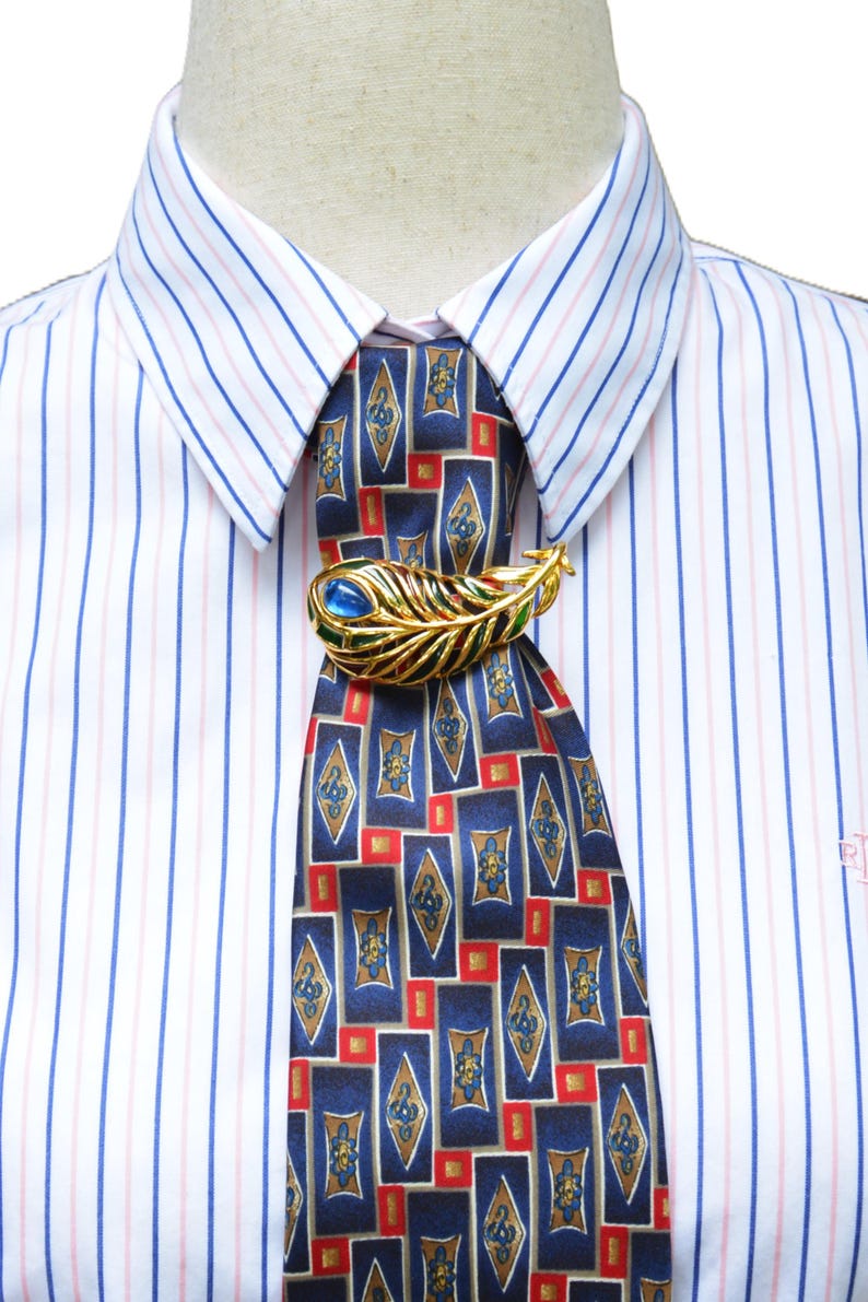 May include: A gold-toned metal brooch shaped like a peacock feather with blue and green enamel accents, pinned to a dark blue necktie featuring a geometric pattern of red and gold rectangles, worn over a white dress shirt with thin blue and pink stripes.