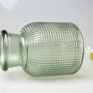 May include: A light green glass vase with a cylindrical body and a short, narrow neck. The exterior surface of the vase features a raised, grid-like textured pattern throughout the main body, while the rim and neck remain smooth and clear.