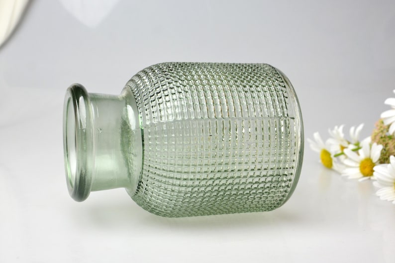 May include: A light green glass vase with a cylindrical body and a short, narrow neck. The exterior surface of the vase features a raised, grid-like textured pattern throughout the main body, while the rim and neck remain smooth and clear.