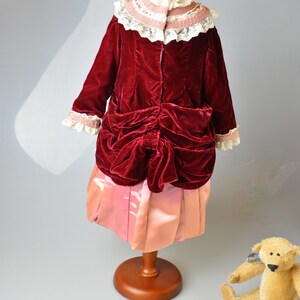 May include: A red velvet dress with a pink satin skirt and a white lace collar. The dress is on a wooden mannequin. A teddy bear is sitting on the floor.