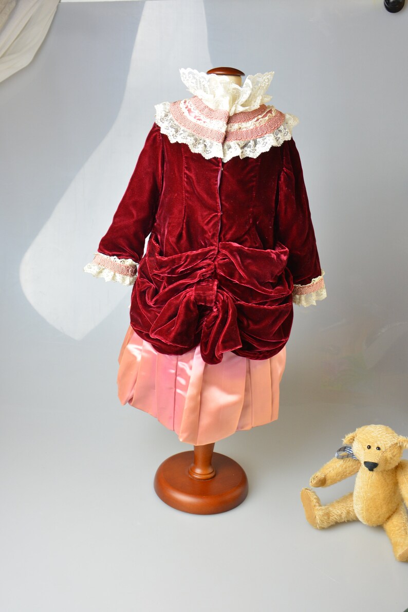 May include: A red velvet dress with a pink satin skirt and a white lace collar. The dress is on a wooden mannequin. A teddy bear is sitting on the floor.