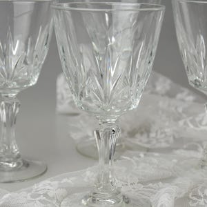 May include: Four clear crystal wine glasses with a detailed, etched pattern. The glasses have a classic design with a stemmed base and a wide bowl. They are arranged on a white lace tablecloth, creating an elegant display.