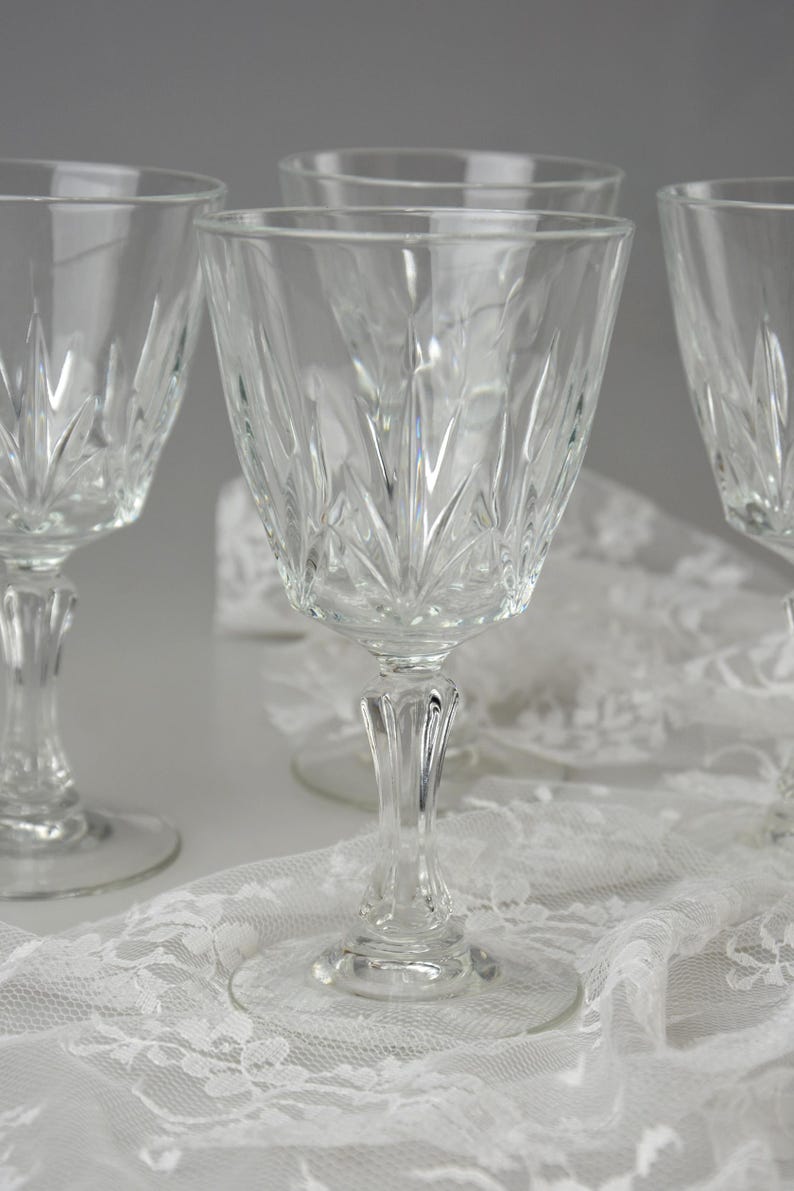 May include: Four clear crystal wine glasses with a detailed, etched pattern. The glasses have a classic design with a stemmed base and a wide bowl. They are arranged on a white lace tablecloth, creating an elegant display.