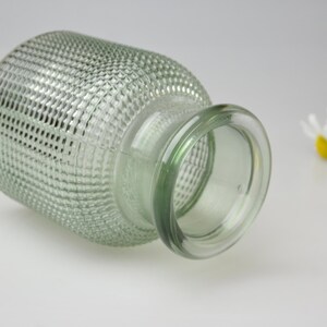 May include: A light green glass vase with a textured, grid-like pattern across its cylindrical body and a smooth, rounded rim. The vase is positioned horizontally, showcasing its translucent material and the small white flowers nearby.