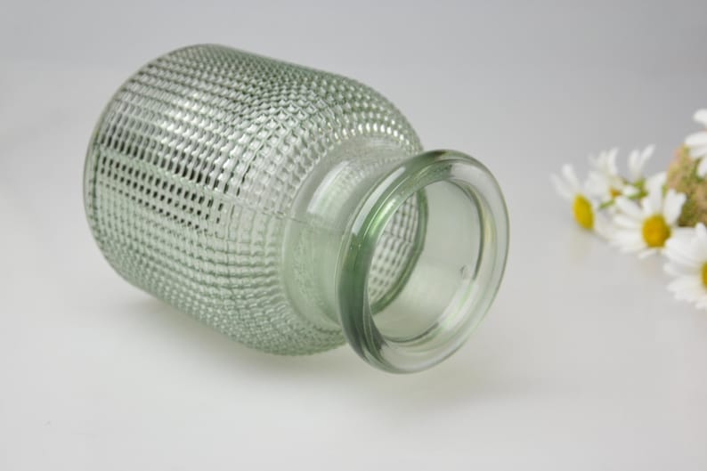 May include: A light green glass vase with a textured, grid-like pattern across its cylindrical body and a smooth, rounded rim. The vase is positioned horizontally, showcasing its translucent material and the small white flowers nearby.