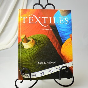 Puede incluir: Un soporte para libros de metal negro con un dise&ntilde;o decorativo de volutas sostiene un libro titulado "Textiles" D&eacute;cima edici&oacute;n por Sara J. Kadolph. La portada del libro presenta una imagen colorida de carretes de hilo.