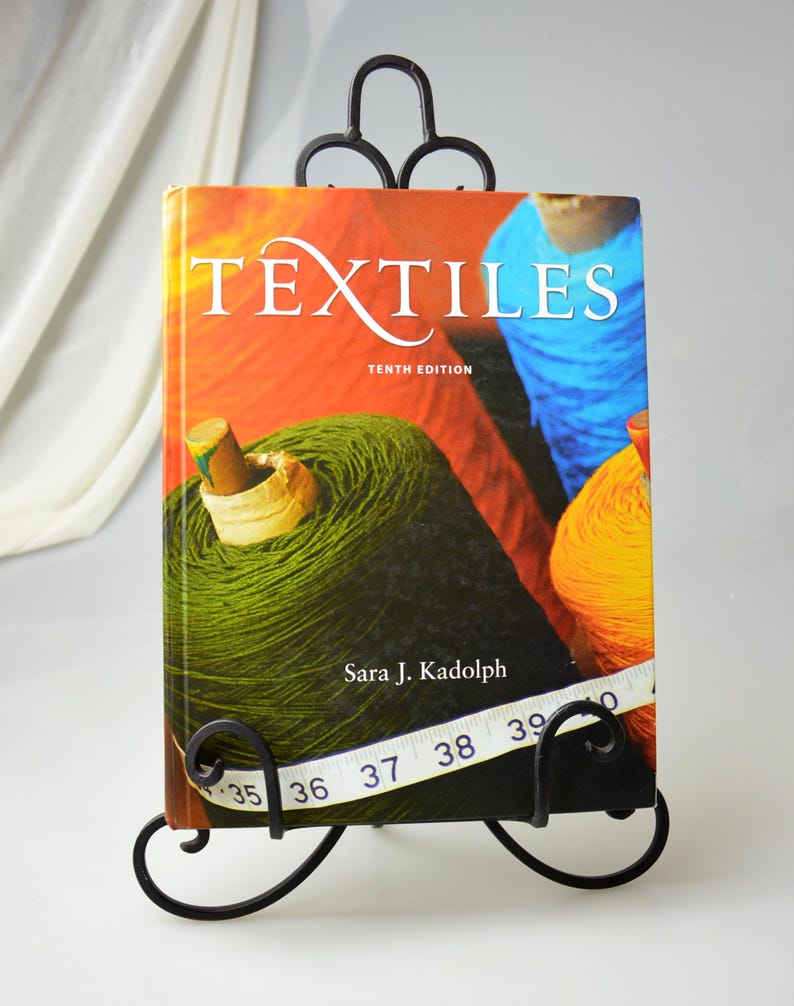 Puede incluir: Un soporte para libros de metal negro con un dise&ntilde;o decorativo de volutas sostiene un libro titulado "Textiles" D&eacute;cima edici&oacute;n por Sara J. Kadolph. La portada del libro presenta una imagen colorida de carretes de hilo.