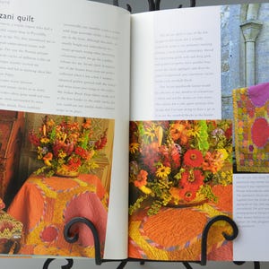 May include: A book spread showcasing a Suzani quilt and floral displays. The quilt, with its patchwork design, features a variety of shapes and colours. The flowers are arranged in a vase on a table covered with an orange tablecloth.
